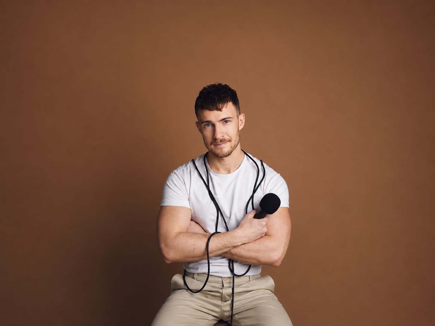 Aidan Wharton, seated against a warm tan studio backdrop, microphone cord crossed over his arms.
