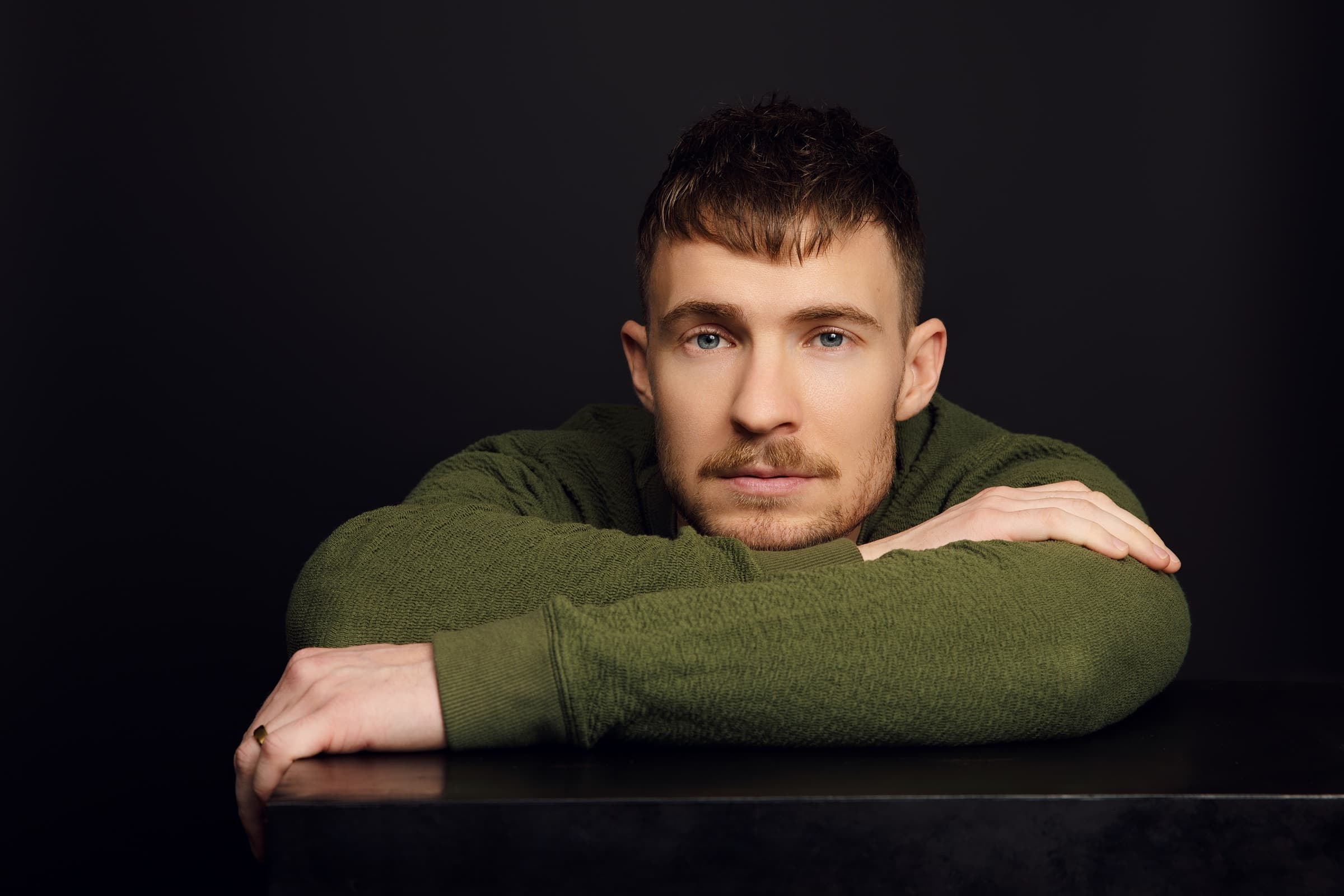 Aidan Wharton in a green sweater, arms folded on a black plinth, against a black backdrop.